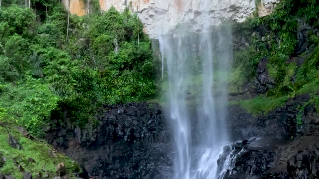 Air terjun yang deras mengalir di atas bebatuan gelap yang dikelilingi oleh tumbuh-tumbuhan hutan hujan tropis hijau yang subur