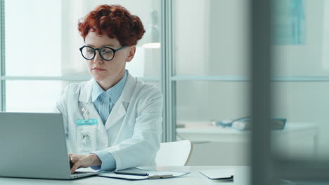 Young-Female-Doctor-Working-on-Laptop-and-Taking-Notes-in-Clinic