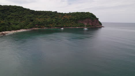 Vuelo-Frontal-De-Drones-Desde-El-Mar-Hasta-Las-Costas-Rocosas-De-Punta-De-Mita,-Con-Barcos-Y-Una-Montaña-Verde-Y-Exuberante-Que-Contrasta-Con-El-Mar-Azul