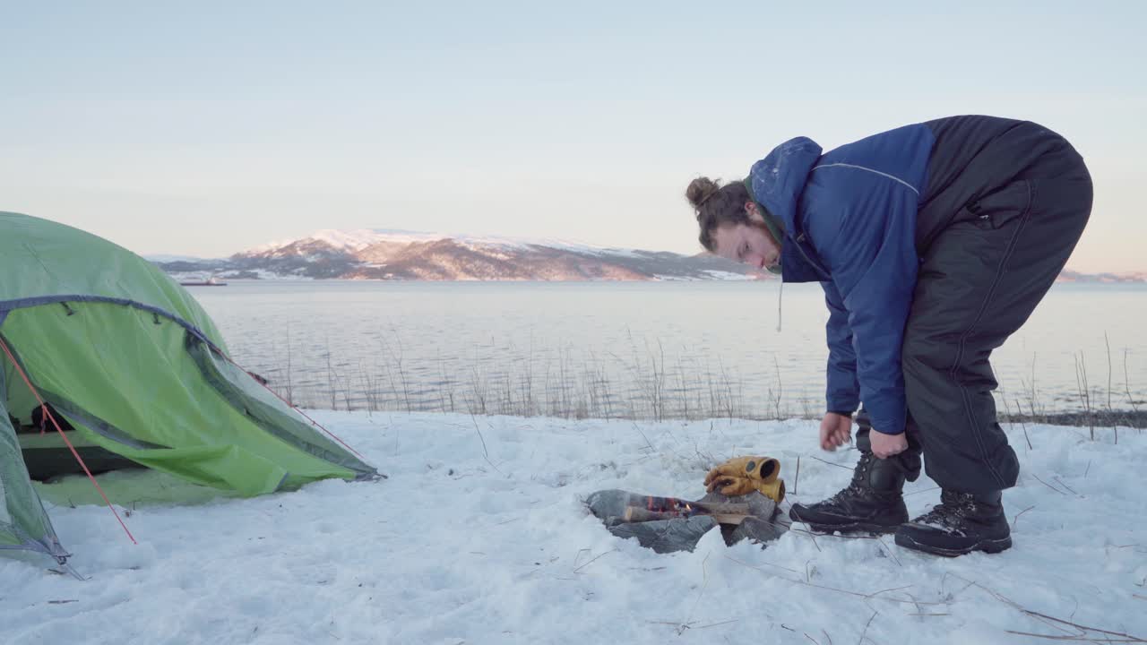 겨울철 텐트 앞 모닥불 근처에서 장화를 묶는 남자