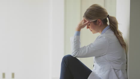 medico femminile stressato che indossa un cappotto bianco seduto sul pavimento nel corridoio dell'ospedale con la testa tra le mani