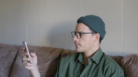 young man with beanie and glasses on couch scrolling on his smartphone
