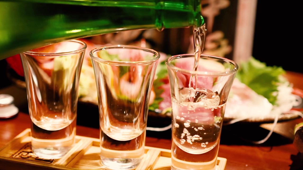 Primo piano di sake trasparente versato da una bottiglia verde in piccoli bicchieri con sashimi fresco.