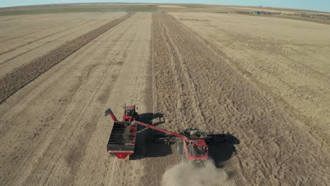 gran máquina agrícola industrial cosecha grano de trigo y transfiere cultivos a un camión rojo en tierras de cultivo planas y expansivas en el campo de ponteix, saskatchewan, canadá, enfoque aéreo superior