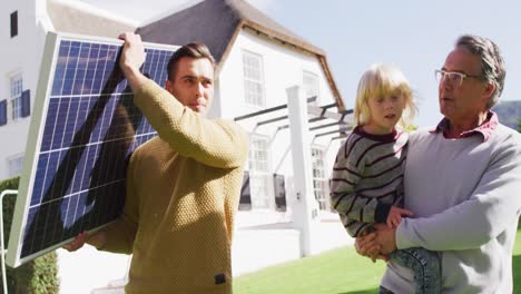 Video-of-happy-caucasian-father-carrying-solar-panel,-grandfather-carrying-grandson-in-sunny-garden