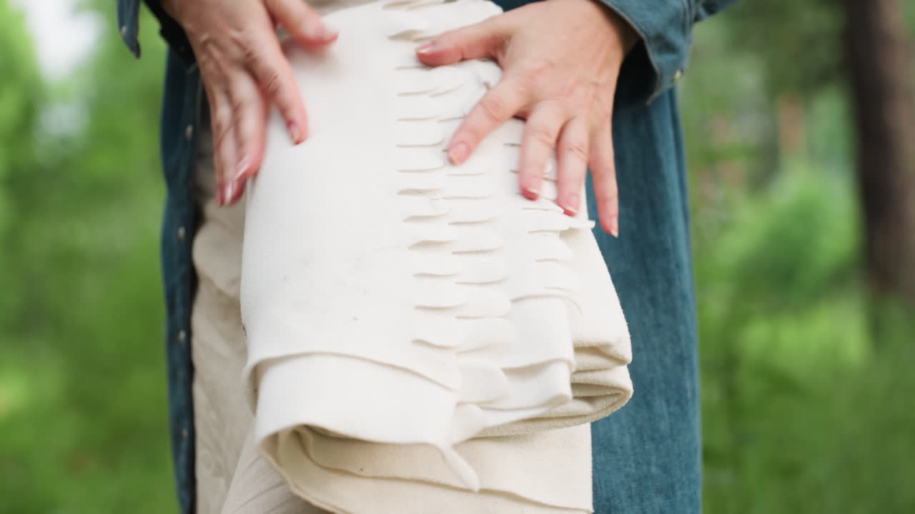 Young mother folding picnic blanket after pleasant outdoor family day in green forest, surrounded by tall trees, calm sunlight, and soft breeze, capturing peaceful end of warm summer moment