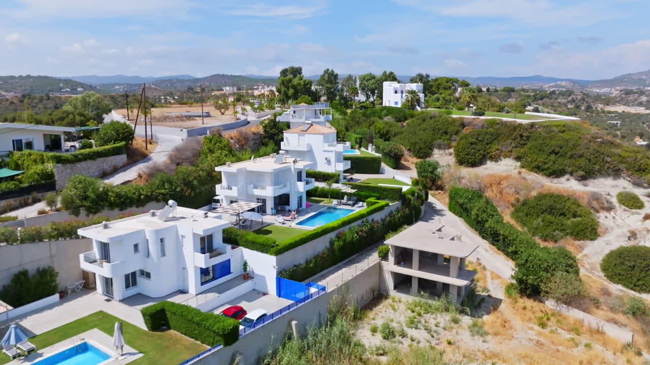 Aerial view passing white luxury holiday homes on a hill, sunny day in Greece