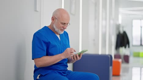 Confident-and-serious-elderly-male-doctor-in-glasses-with-a-gray-beard-and-blue-uniform-sits-on-social-media-during-his-break-in-the-corridor-of-a-modern-clinic