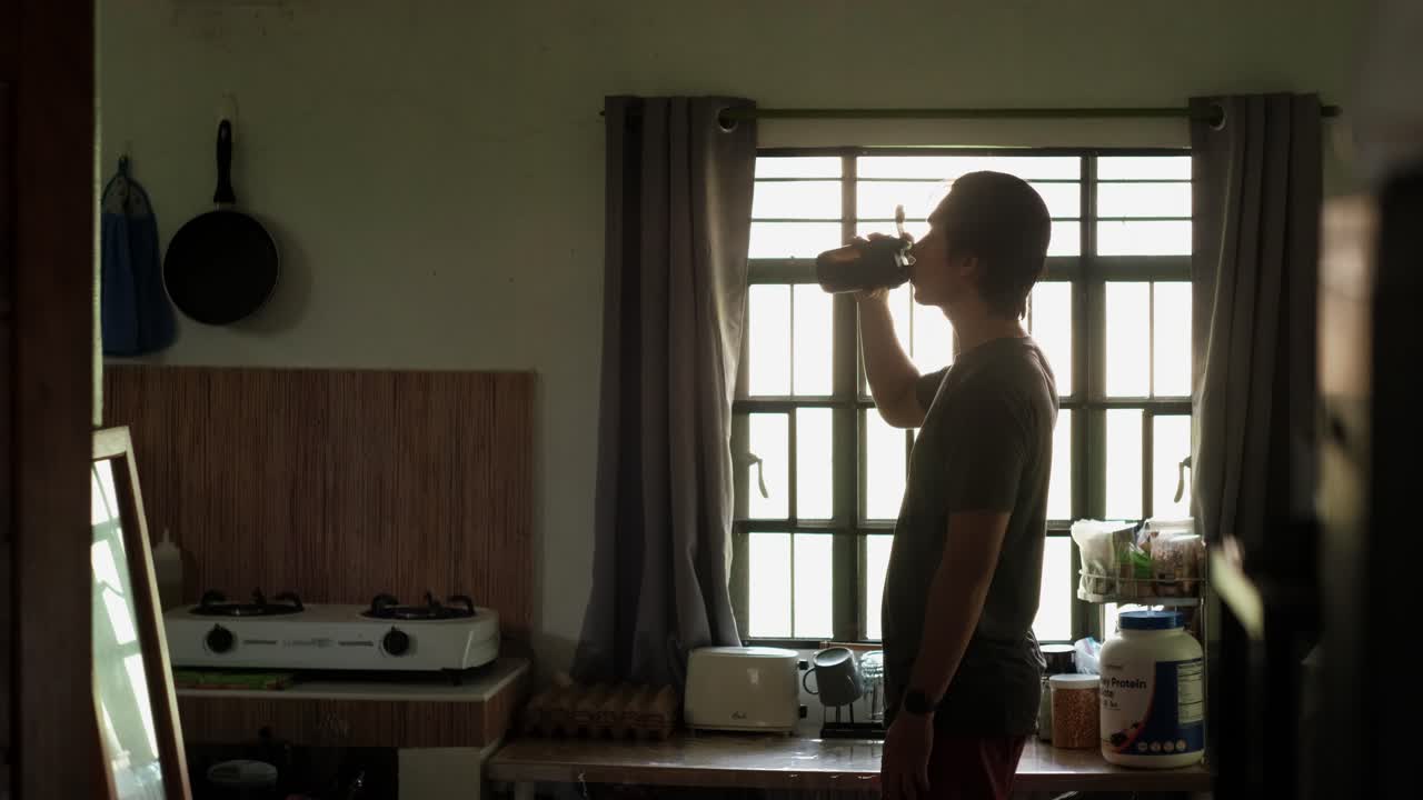 Static Medium shot of a Young Man Shaking and Drinking from Protein Shaker Bottle at Home Kitchen during sunrise, post-workout