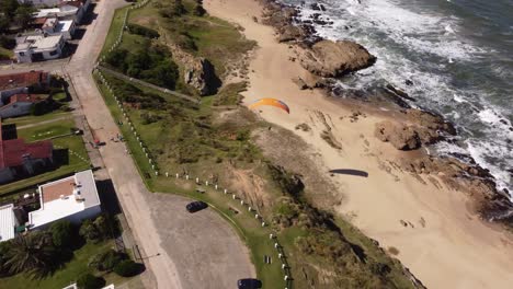 Toma-Aérea-De-Parapente-Volando-Sobre-Una-Playa-Rocosa-En-Un-Día-Soleado-4k