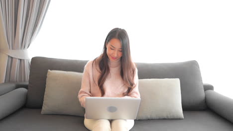 Eine-Attraktive-Frau-In-Einem-Pullover-Und-Weißen-Jeans-Sitzt-Auf-Der-Couch,-Während-Sie-An-Ihrem-Laptop-Arbeitet