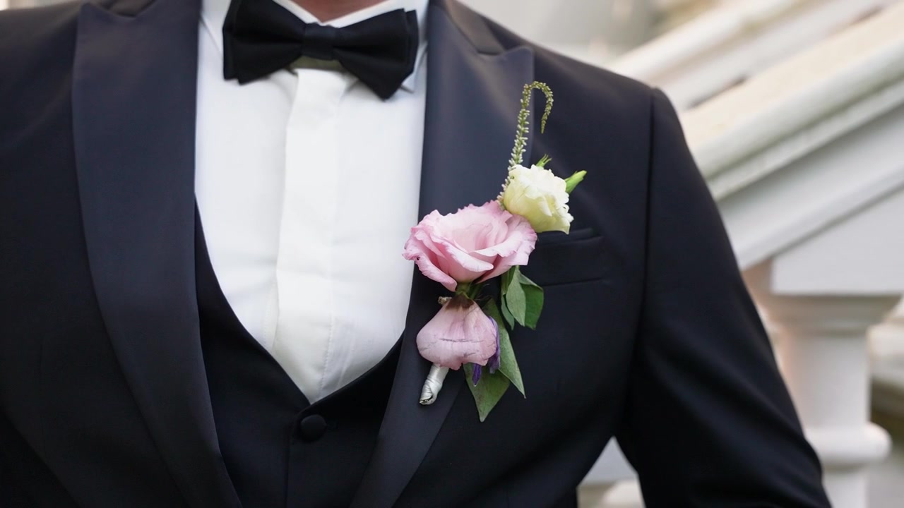 Primo piano cinematografico di uno sposo in un classico smoking nero con una boutonnière pastello, in piedi davanti a una scalinata ornata. Messa a fuoco nitida, luce naturale, eleganza nuziale, dettaglio intimo.
