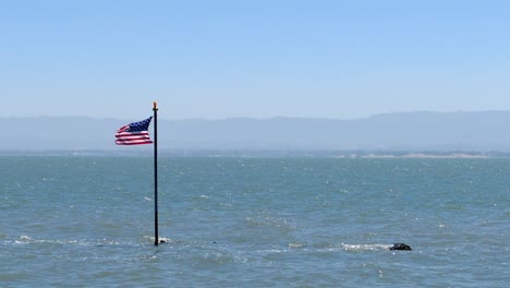 Stars-and-stripes-blowing-in-the-wind-on-flag-pole-coming-out-the-ocean-with-waves-and-mountains-on-the-horizon