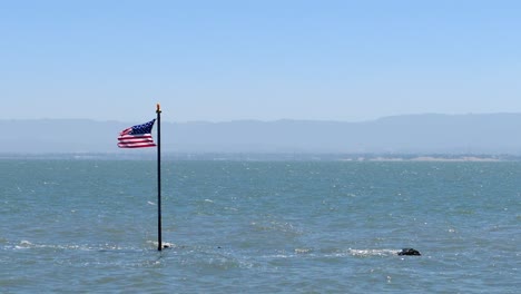 Sterne-Und-Streifen-Wehen-Im-Wind-Auf-Einem-Fahnenmast,-Der-Aus-Dem-Meer-Kommt,-Mit-Wellen-Und-Bergen-Am-Horizont