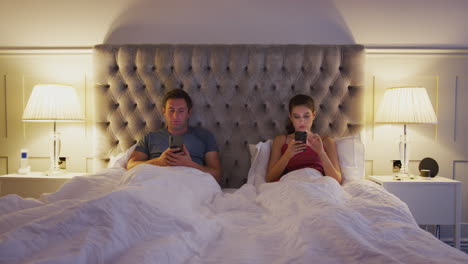 couple lying in bed checking mobile phones whilst ignoring each other