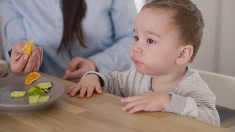 unrecognizable mother feeding her little baby boy with segments of clementine while sitting together next to the table in living room