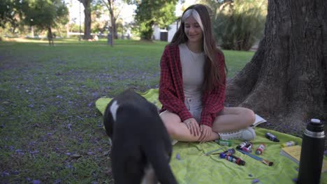 Pretty-young-woman-caressing-her-dog-sitting-under-tree-surrounded-by-paintings-and-book-at-sunset