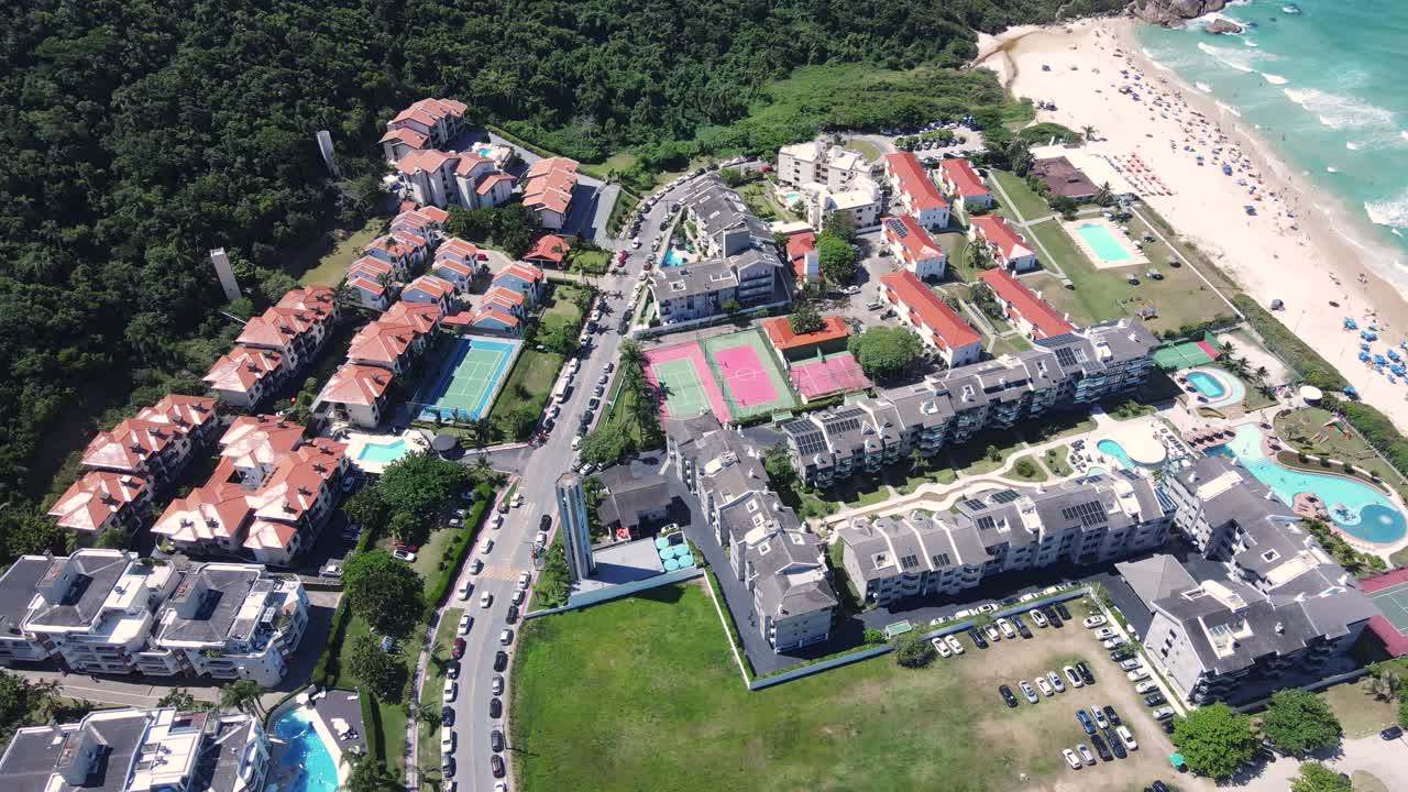 Aerial drone shot of a beautiful turquoise blue sea beach with mountains covered in vegetation near the coast with many summer condominiums amid the Atlantic Forest and Ocean in Florianopolis