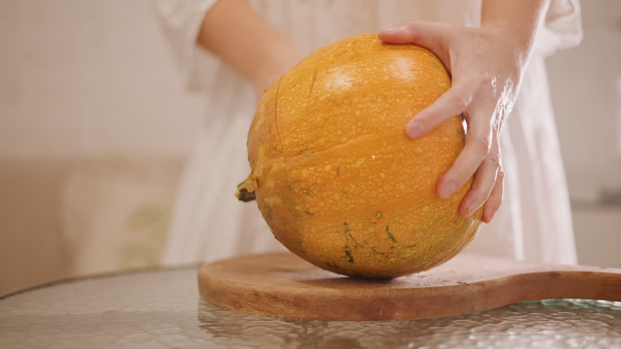 Manos cortando una calabaza entera en una tabla; primer plano de manos cortando una calabaza naranja grande en una tabla de cortar de madera en una cocina luminosa y acogedora, con textura rústica, preparando el relleno para pastel de calabaza, bajo una luz cálida y suave.