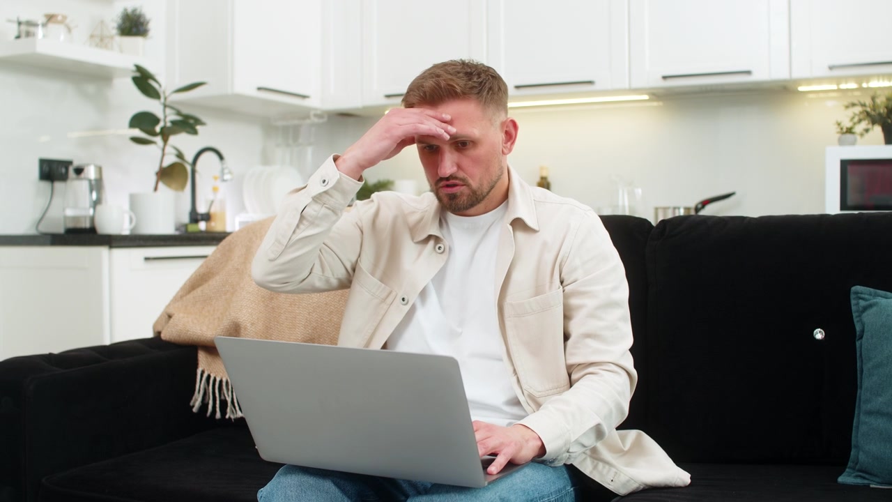 Young man sees defeat on laptop touches forehead sighs and clicks retry after bad news on screen sad