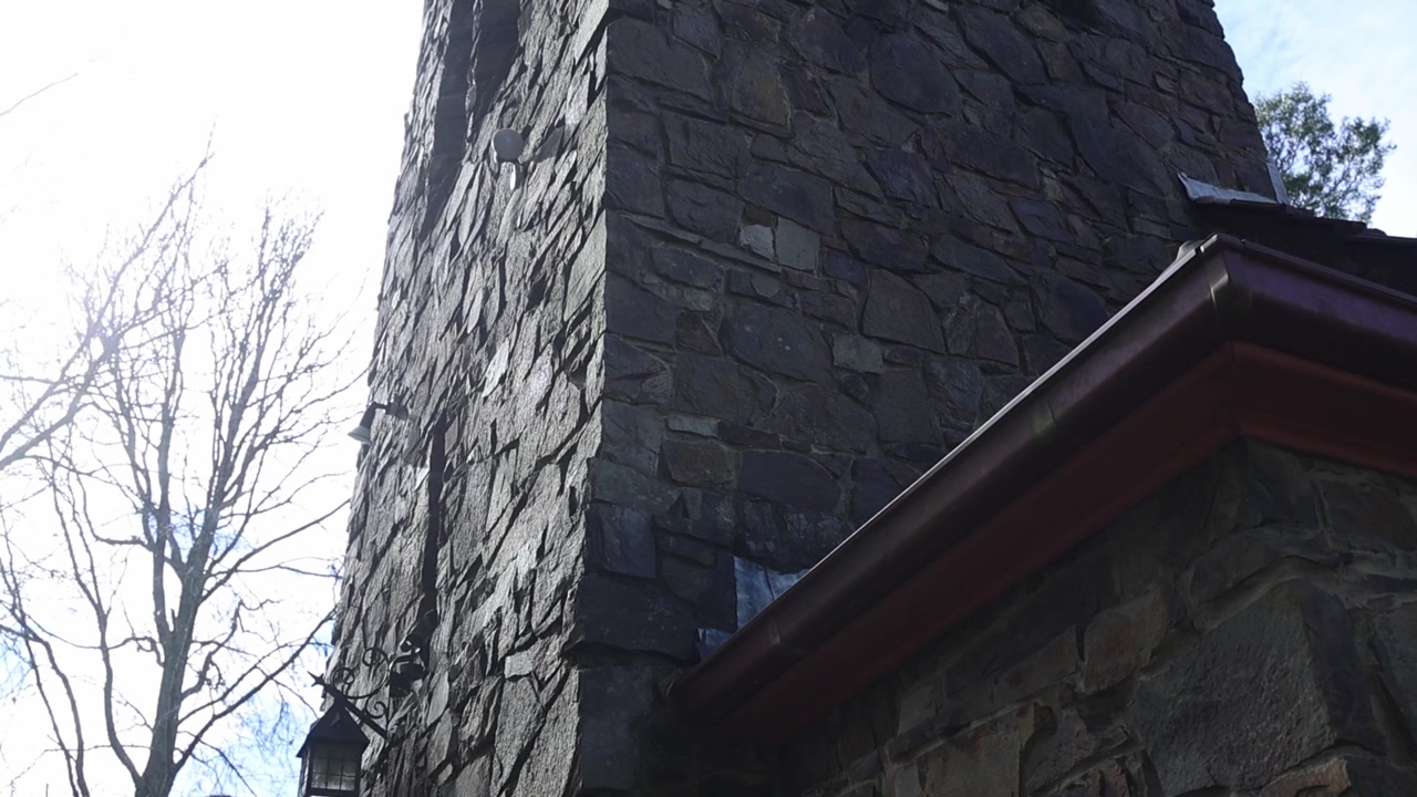 Low-angle tracking shot of old stone church tower in Olinda, Melbourne, with textured masonry, dramatic shadows, and bright natural daylight highlighting rustic details. No people visible.