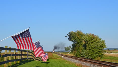 a view of a line of gently waving american flags on a fence by farmlands as a steam passenger train blowing smoke approaches during the golden hour