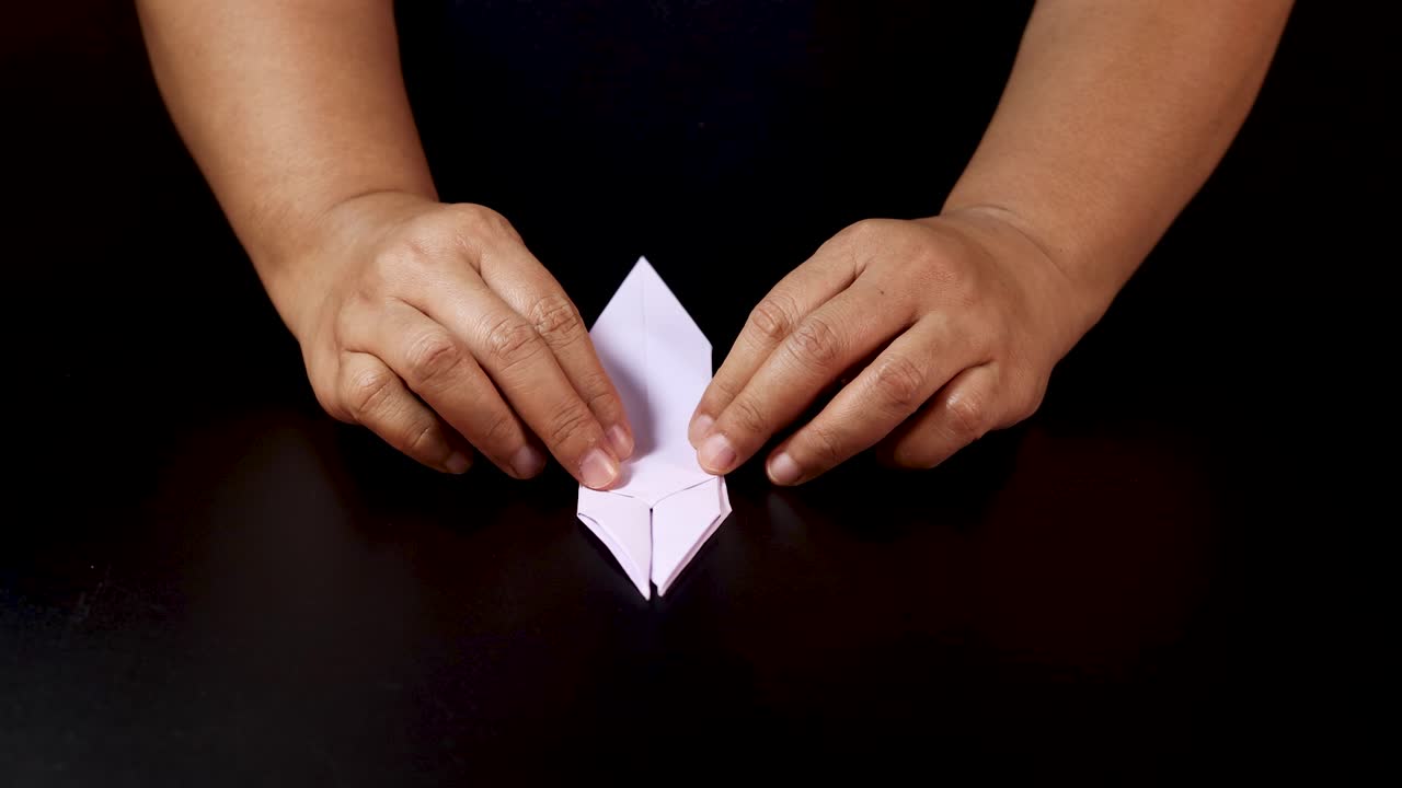 Adult hands folding pink origami bird on black surface, overhead view, even studio lighting
