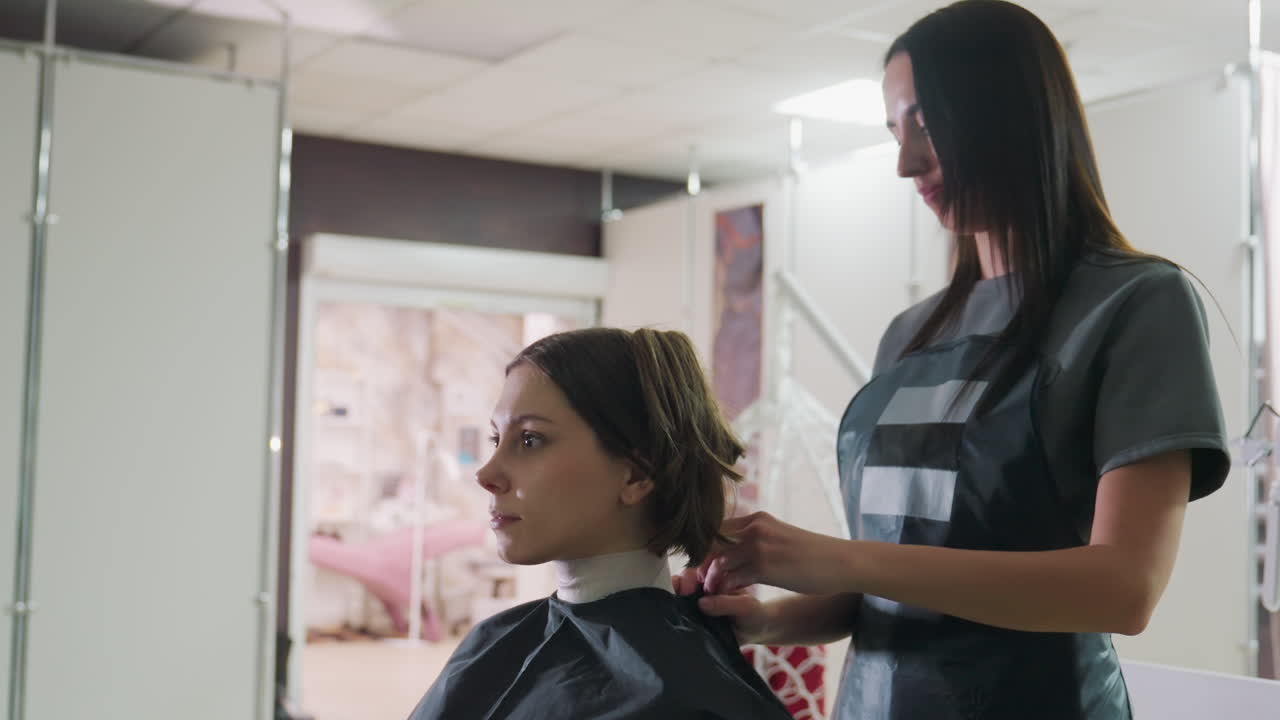 En klient sidder i en salonstol under en opmærksom frisørkonsultation og justering af kappe, blødt dagslys gennem vinduet, spejlbillede, rolig professionel atmosfære, hårforberedelse til klipning