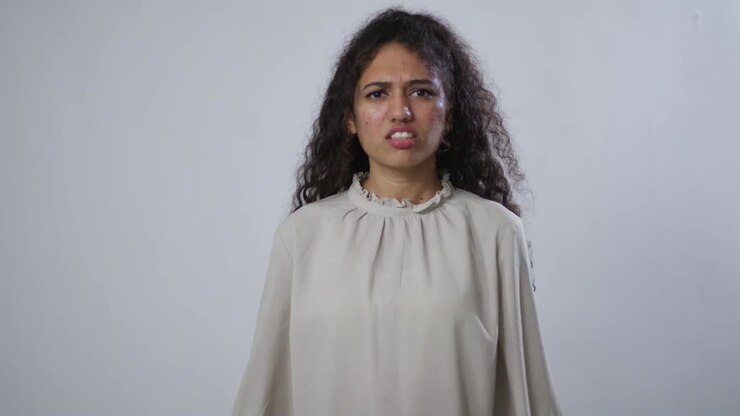 A South Asian woman with curly hair, in a light shirt, nods her head to signify "no", with a disgusted or disapproving face