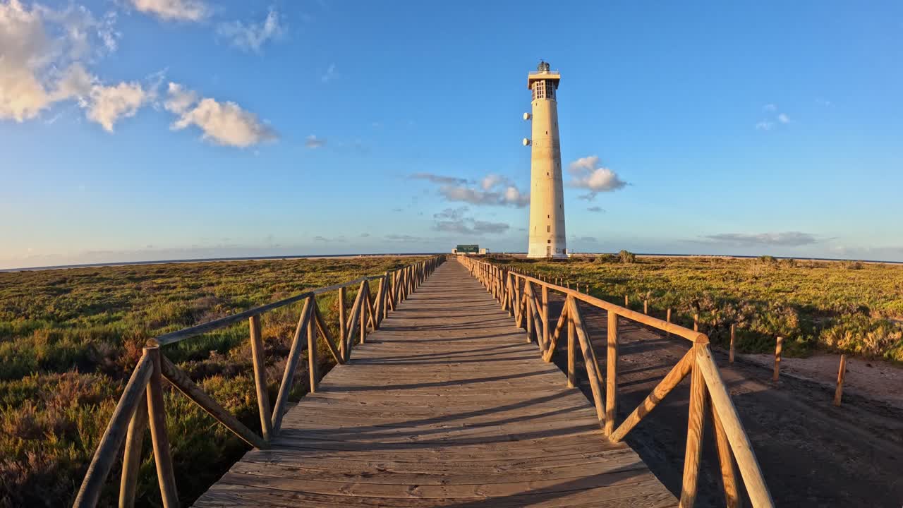 Walking on a wooden boardwalk stretches across a green coastal landscape, leading to a tall lighthouse under a golden sunset sky.