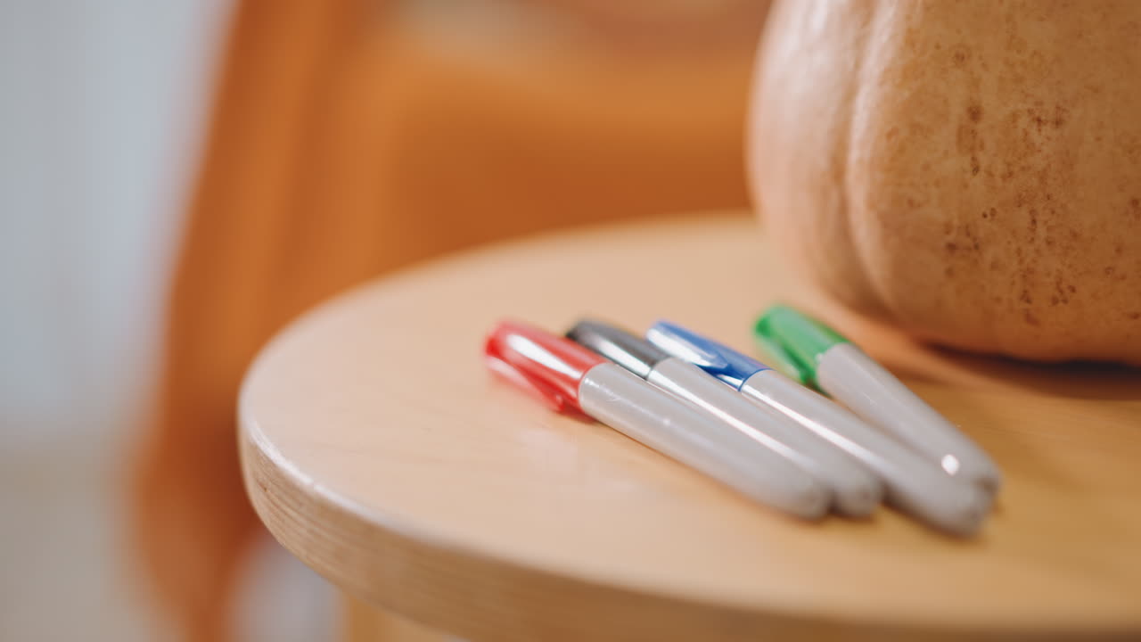 Closeup pumpkin and markers on table, warm orange backdrop, wooden stool edge, shallow depth of field, soft bokeh, cozy autumn craft scene with stationery ready for seasonal decoration