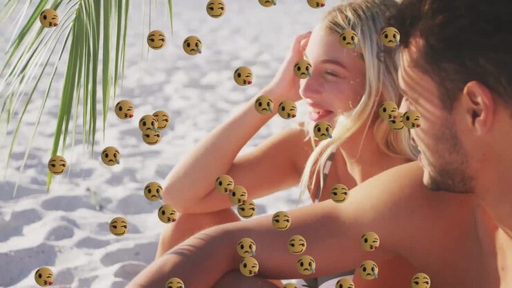 Emoji multiple di volti che fluttuano sullo sfondo di una coppia caucasica che sorride mentre è seduta in spiaggia