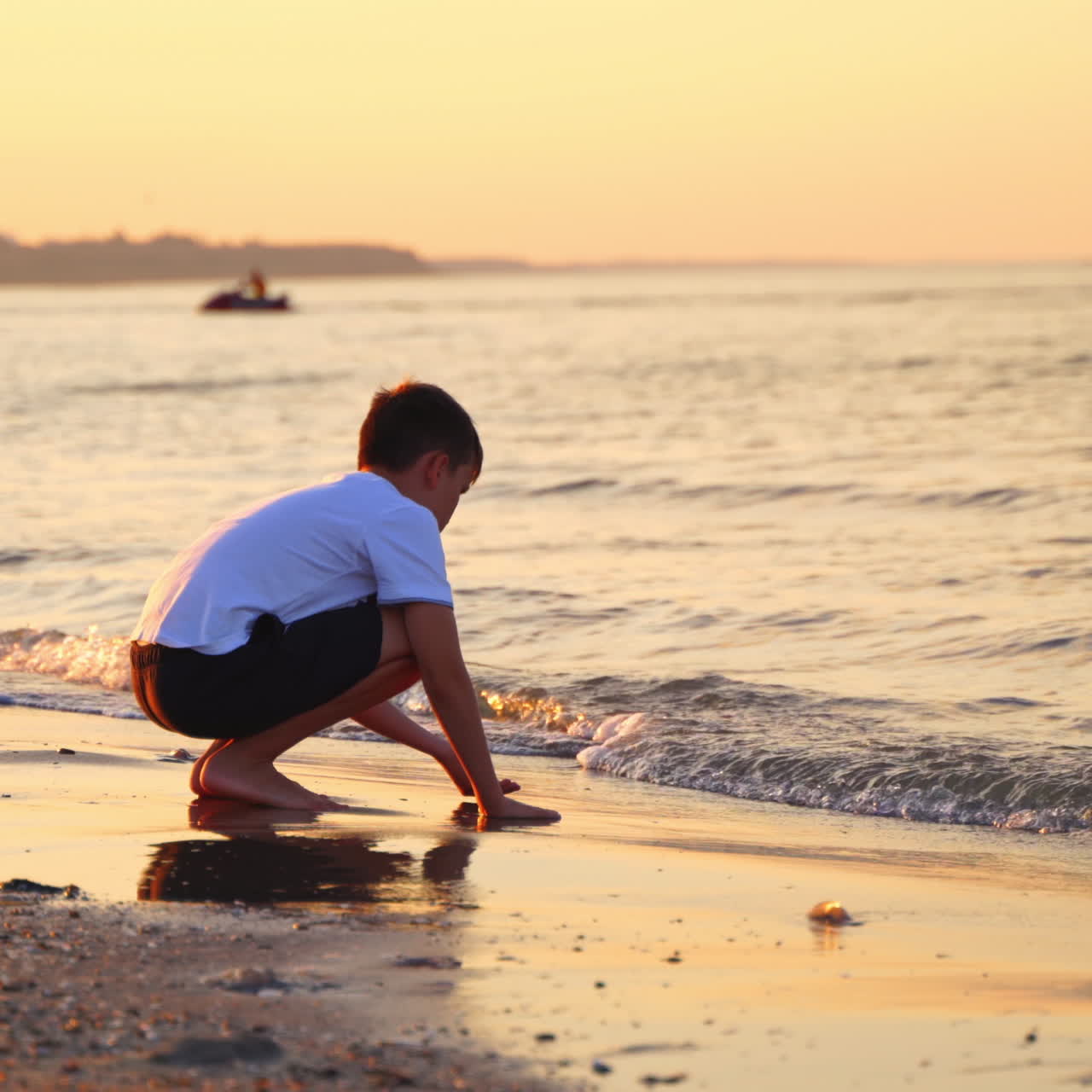 Boy plays in sea water in the evening. Side view of a boy in white t-shirt and shorts playing on sea shore at sunset.