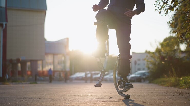Person with short brown hair wearing gray hoodie and dark gray pants jumping on spring stilts on paved road in residential area surrounded by houses, showcasing unique motion and urban lifestyle