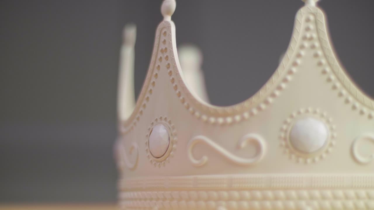 Close up of a beige crown spinning on a pedestal with ornaments and white pearl jewels with peaks jagged out of plastic. With a neutral grey background. Shot in 4K.