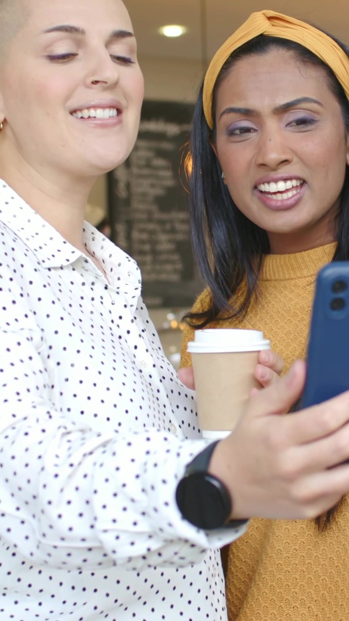 Vertical video: Adjusting blue phone, mustard woman showing friend with cup at cafe seeing selfie
