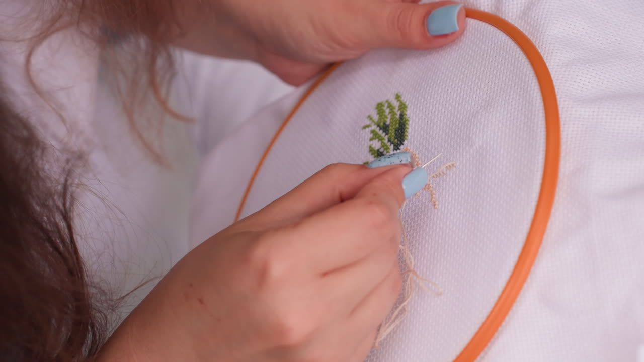 Close up of stylist with painted blue nails delicately embroidering fabric in orange hoop, focusing on green thread pattern, showing artistic patience, precision, and dedication