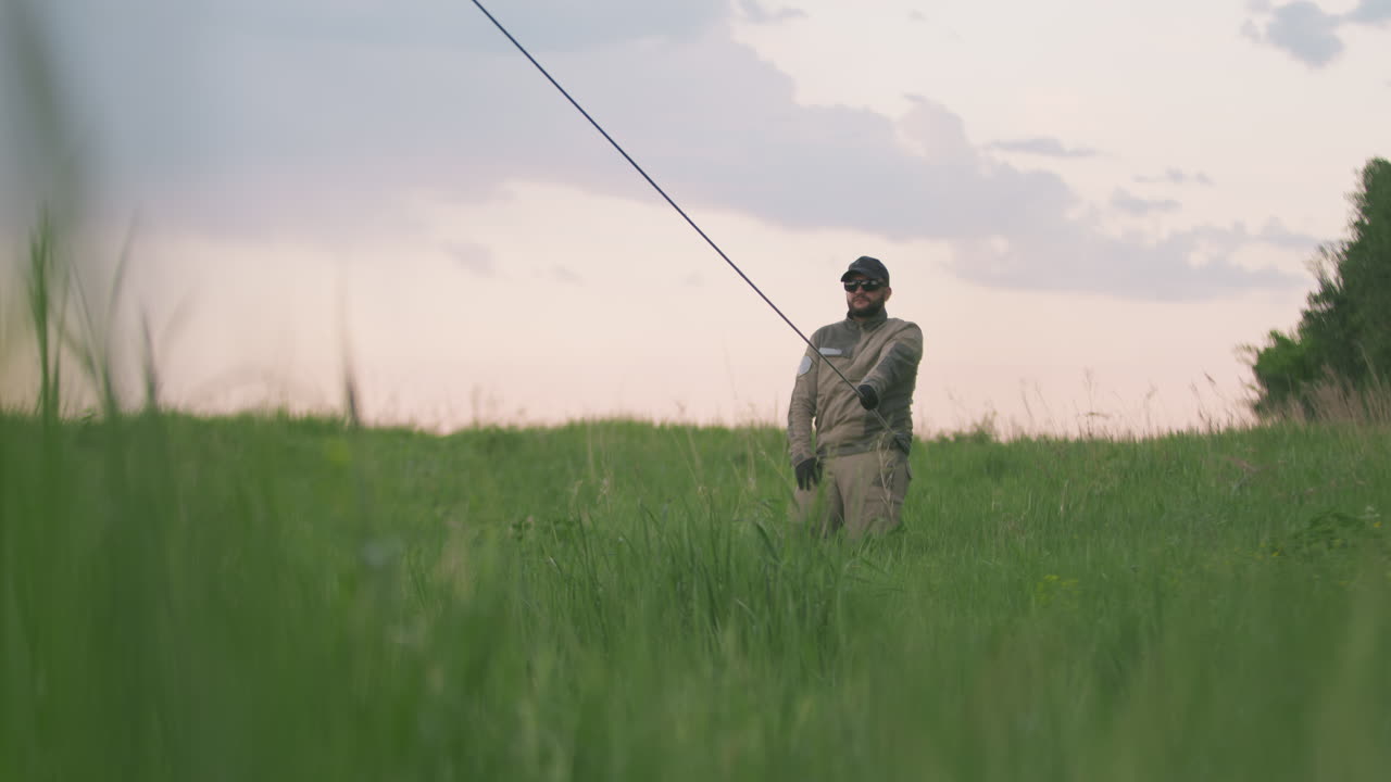 Man in dark shades gripping tether rope and stepping forward through tall green grass as part of hot air balloon setup under soft pastel evening sky, distant tree line and fading light