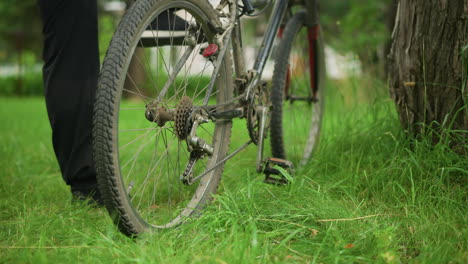 leg view of someone in black trousers and sneakers walking up to a parked bicycle in grassy field, he adjust the pedal, lift the back tires, and pedal, causing the tires to rotate briefly