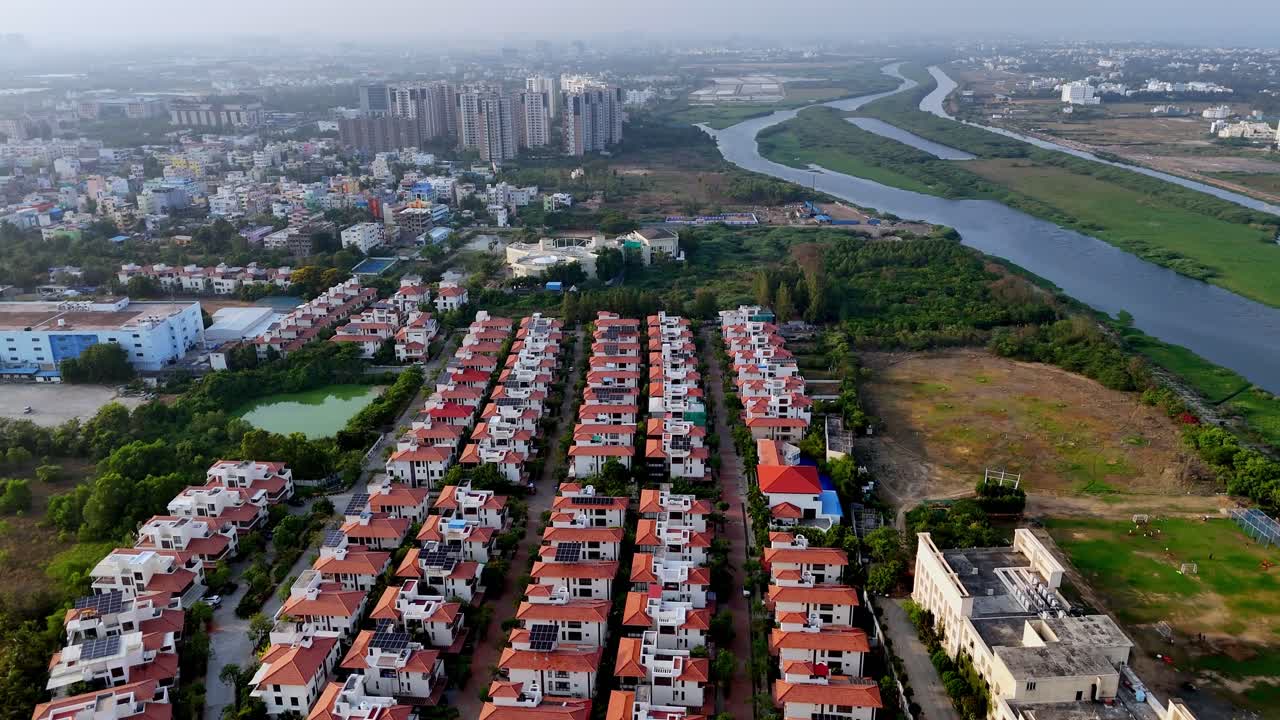 High-angle aerial view of a modern residential area in India, featuring rows of neatly arranged gated community homes with red-tiled roofs. a winding river or canal system flows through a lush green
