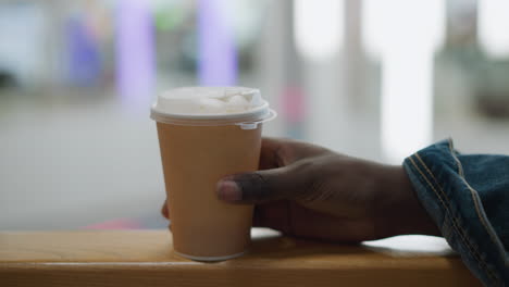 close-up of hand holding paper cup of coffee, with focus on lid and cup details, set against blurred background, moment of calm and reflection in modern setting