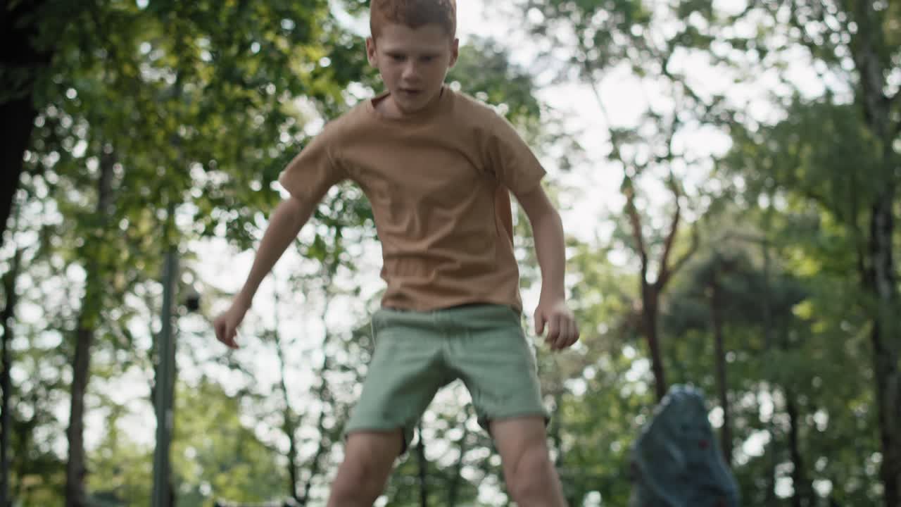 백인 홍색 소년이 놀이터에서 트램폴린에 뛰어다닌다.