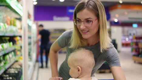 Young-mother-is-choosing-cabbage-and-other-vegetables,-while-her-little-child-is-sitting-in-a-grocery-cart.-Family-shopping-in