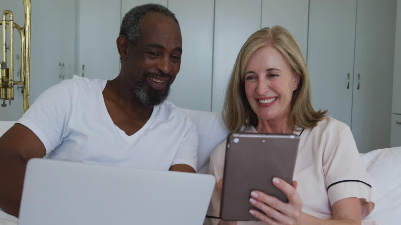 Diverse senior couple sitting in bed using laptop and tablet talking and lsmiling
