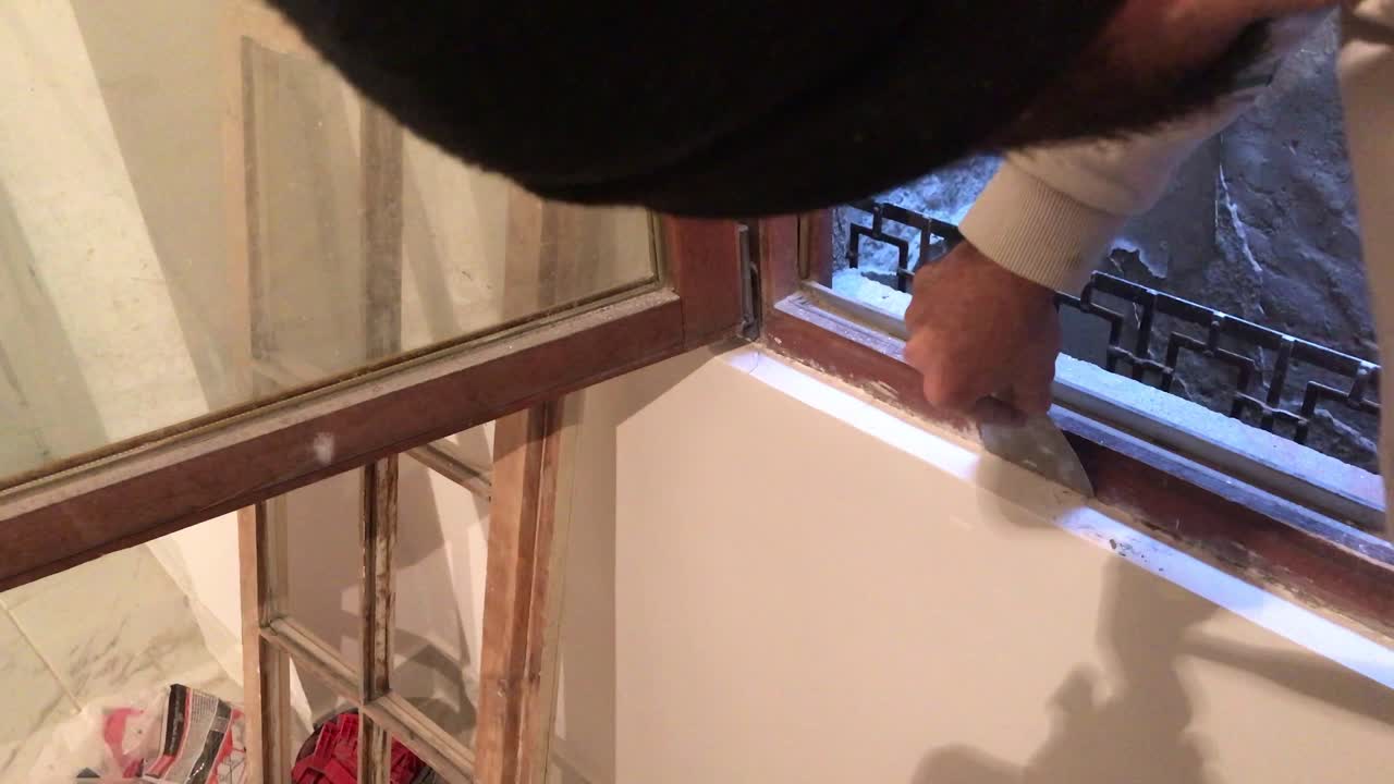 Un giovane che rimuove l'eccesso di intonaco con una spatola attorno a un telaio di finestra in legno in 4k.