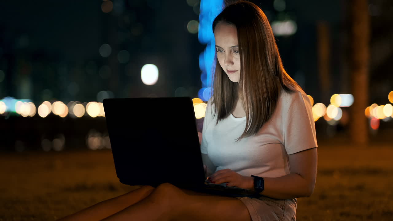 una mujer desarrolladora de software se sienta en un parque en una noche de verano en la ciudad y escribe código mirando la pantalla de una computadora portátil. imprime el artículo en una computadora portable. blogger responde a las publicaciones