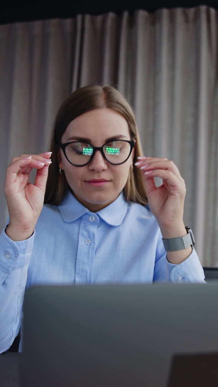 Female employee working on her laptop in the office. Lady takes off glasses, closes laptop and pushes it away looking in the window. Vertical video