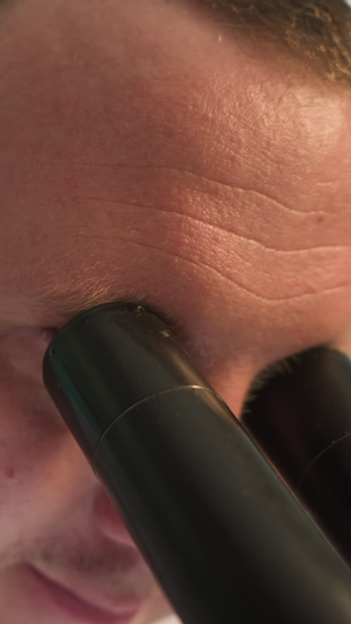 백색 실험실 코트를 입은 기술자가 현미경으로 무언가를 검사하는 근접한 모습, 기술자의 얼굴은 약간 흐릿하며, 전면에 있는 현미경에 초점을 맞추고 있습니다.