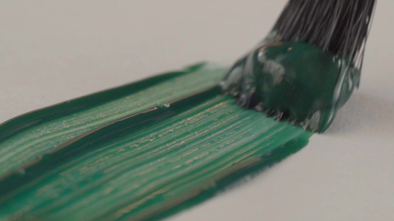 Primo piano macro di un pennello che applica un rivestimento acrilico verde su una superficie bianca. Setole morbide stendono uniformemente il colore denso, rivelando una texture lucida e un movimento fluido