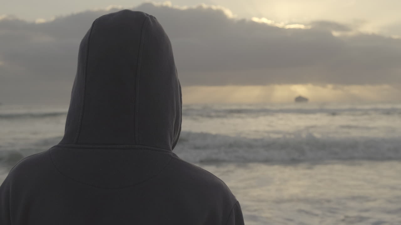 Rear view carefree hooded young adult male watching ship on the distant sunset horizon